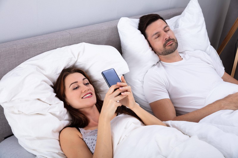 Woman Using Smartphone While Her Husband Sleeping On Bed